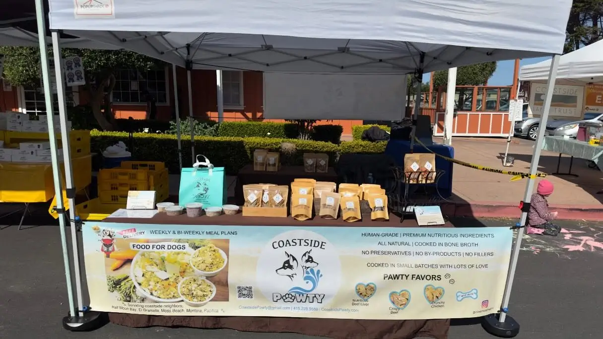 A vendor's booth at a market featuring various dog treats and products, with a sign that reads "COASTSIDE PAWNY" and a display of items including bags of treats and a menu for dog food.